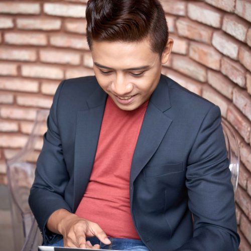 smiling-young-asian-man-sitting-brick-wall-using-tablet_11zon